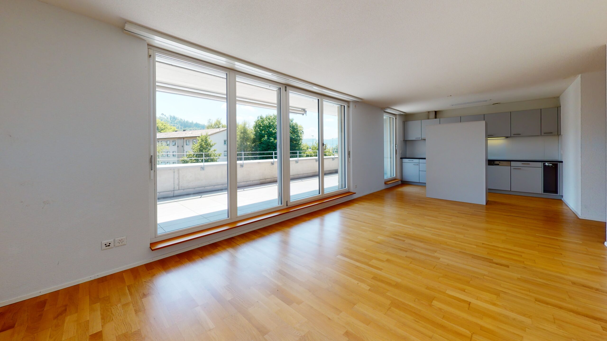 Geräumiger, sonnendurchfluteter Raum mit polierten Holzböden und grossen, raumhohen Fenstern mit Blick auf einen Balkon. Im Hintergrund ist eine moderne Küche mit eleganten Schränken zu sehen.