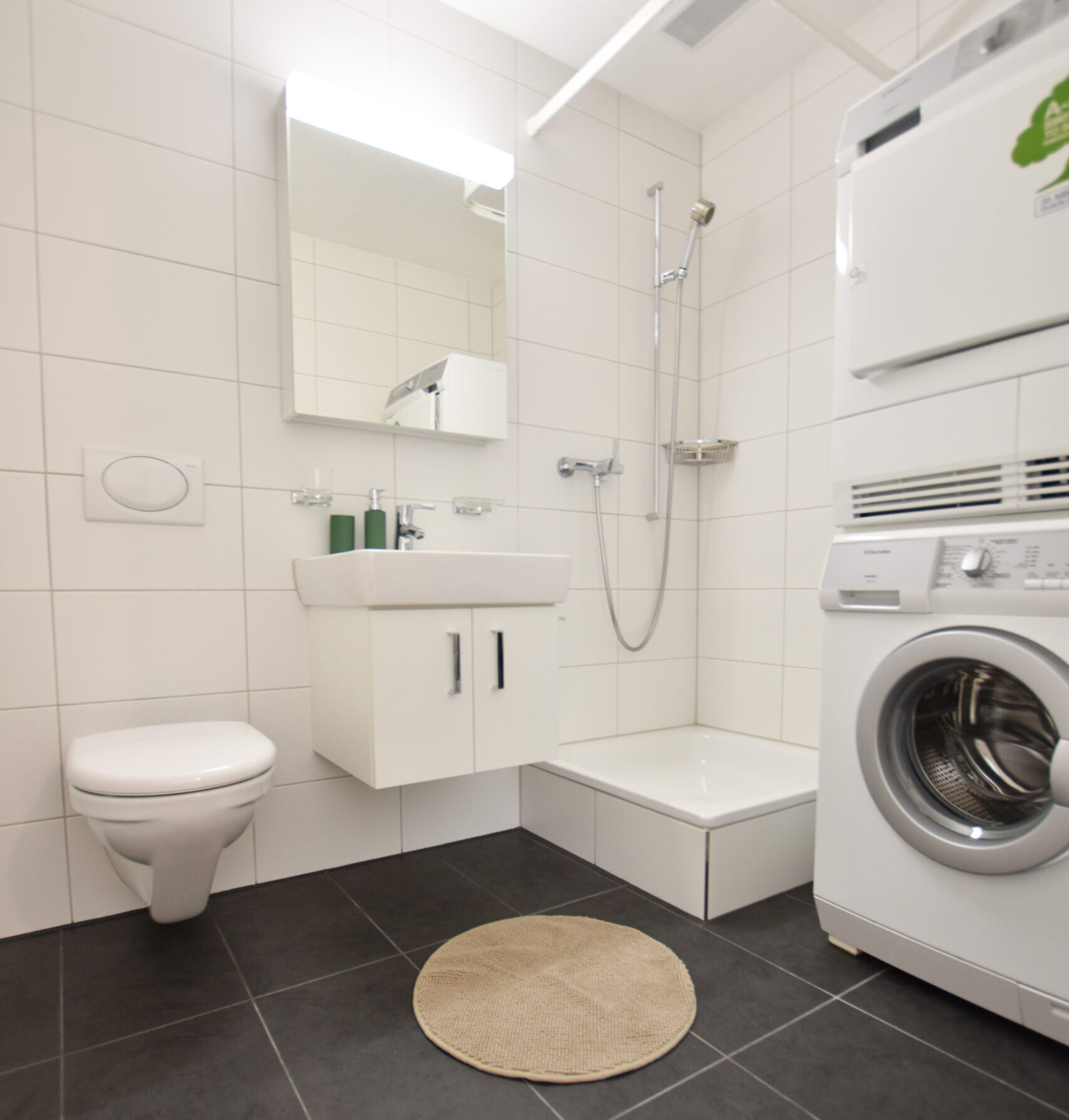 Une salle de bain moderne avec des carreaux blancs. Elle dispose d'un WC suspendu, d'un petit lavabo surmonté d'un miroir et d'un espace douche. À côté de la douche se trouve une unité de machine à laver et de sèche-linge empilée l'une sur l'autre. Un tapis rond beige est posé sur le sol carrelé foncé.