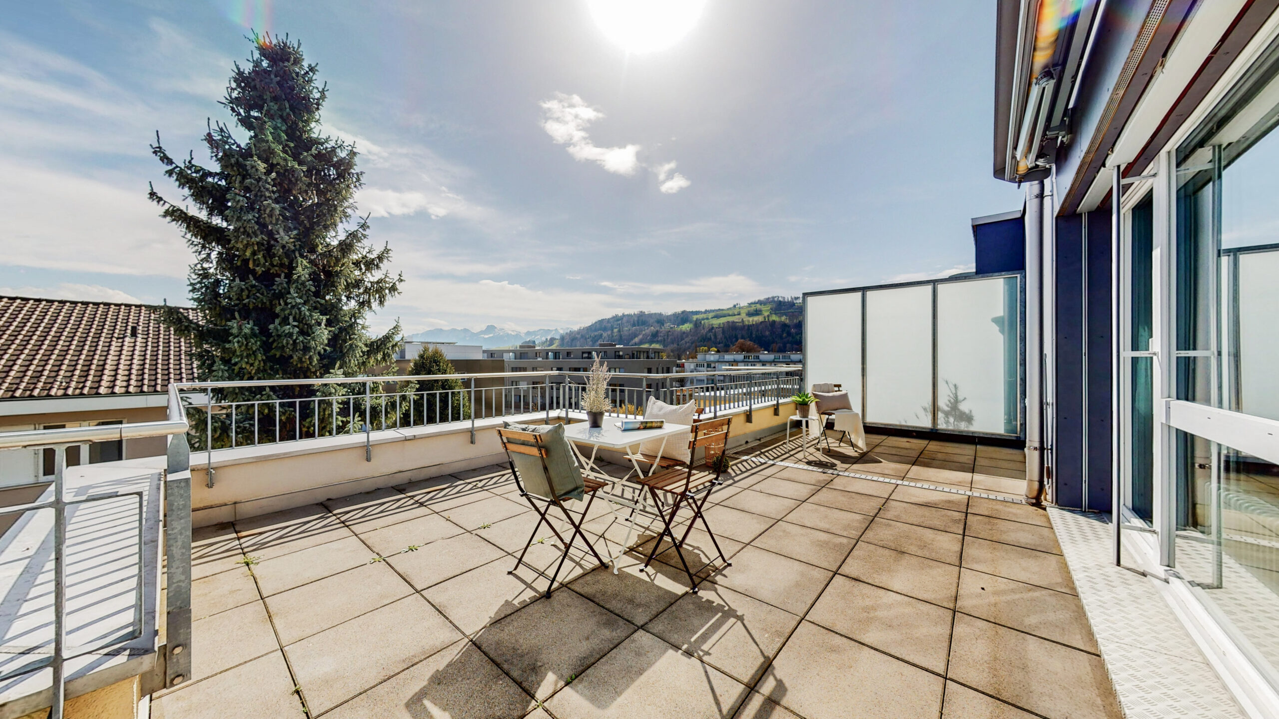 Une terrasse ensoleillée sur le toit, avec une petite table et deux chaises, offre une vue sur un paysage pittoresque de collines et d'arbres. La terrasse dispose d'une balustrade et de grandes portes vitrées sur le côté droit. Au loin, on aperçoit un grand arbre vert.