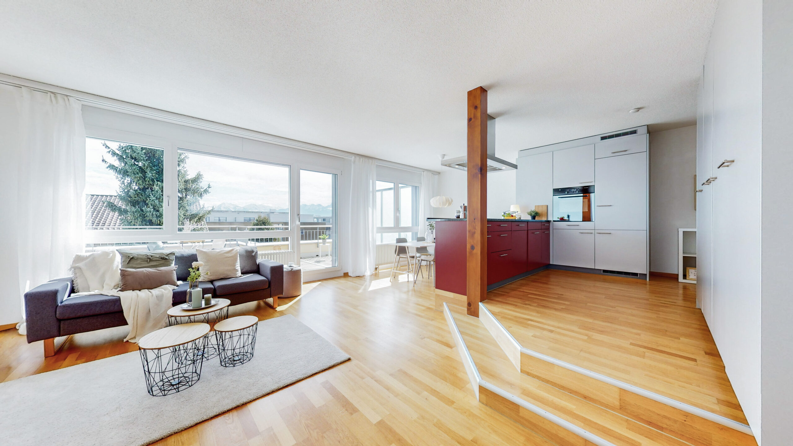 Un espace ouvert moderne pour le salon et la cuisine avec un sol en bois. Le salon dispose d'un canapé gris, de tables basses rondes et d'une grande fenêtre qui donne sur un balcon. La cuisine a des armoires blanches et une table à manger avec des chaises. La pièce est inondée de lumière naturelle.