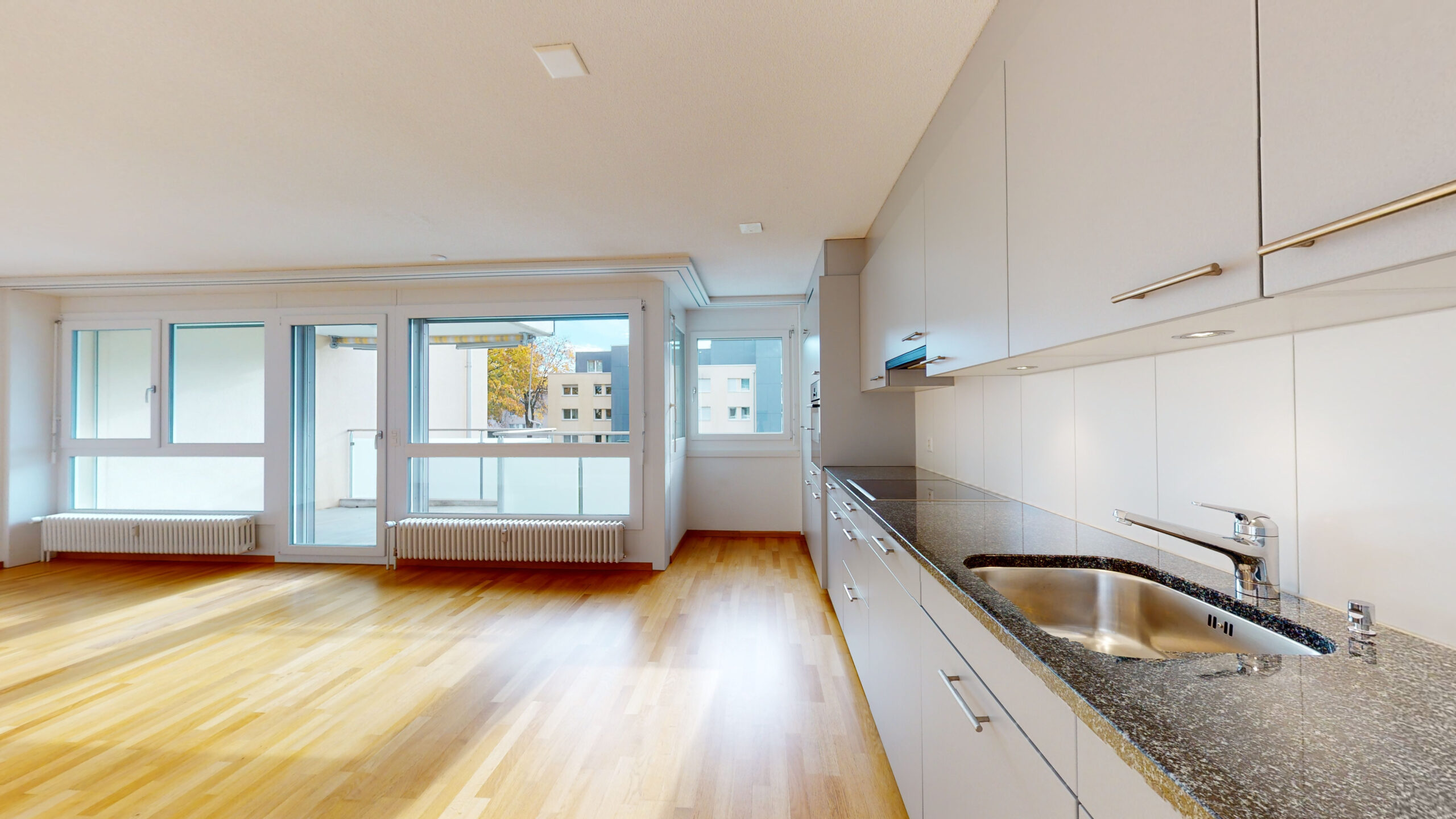 Intérieur d'appartement lumineux et moderne avec un plan ouvert et un sol en bois. La cuisine sur la droite a des armoires blanches et un plan de travail sombre avec évier. De grandes fenêtres sur la gauche apportent la lumière du jour et offrent une vue sur les bâtiments environnants.