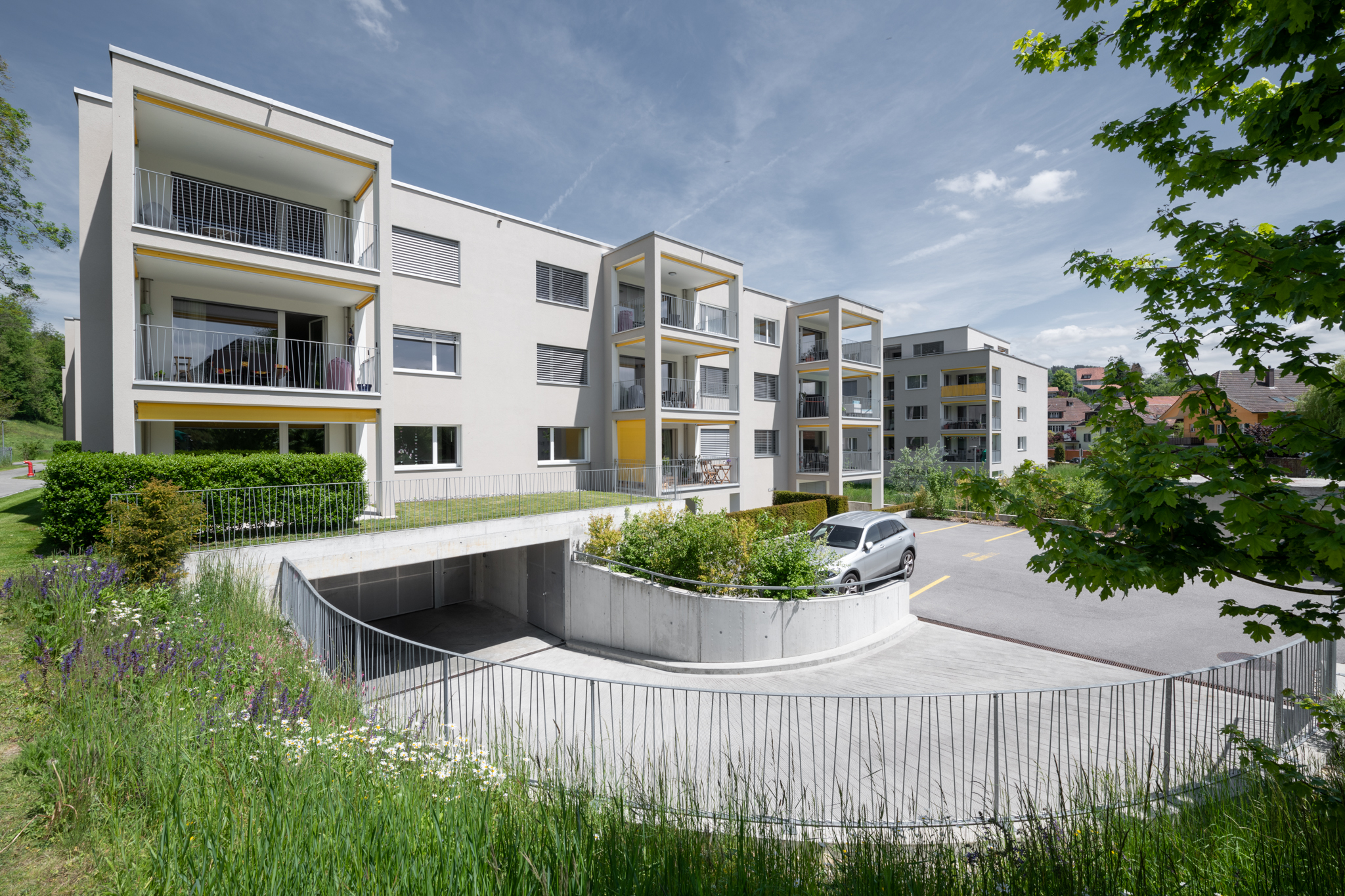 Immeuble d'appartements moderne de trois étages avec balcons et entrée de garage. Une voiture est garée dans l'allée, au premier plan on peut voir des espaces verts et une clôture incurvée. Le ciel est partiellement nuageux.