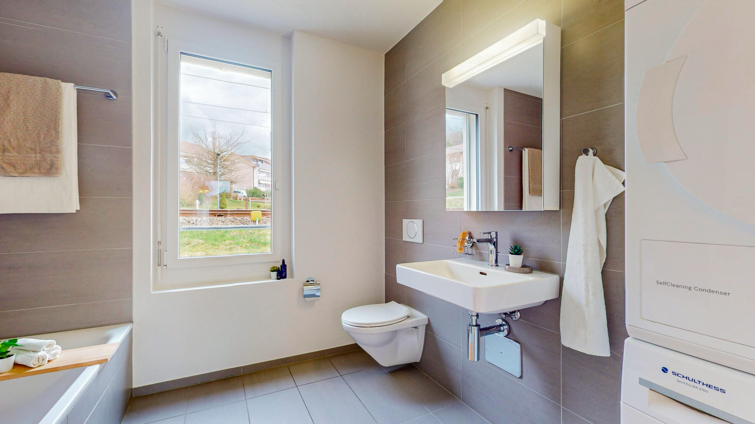 Une salle de bain moderne dans des tons neutres dispose d'une baignoire, d'un lavabo mural et de toilettes. Une grande fenêtre permet à la pièce d'être inondée de lumière naturelle. Une serviette est accrochée au mur et un miroir se trouve au-dessus du lavabo. Une machine à laver est installée dans un coin.