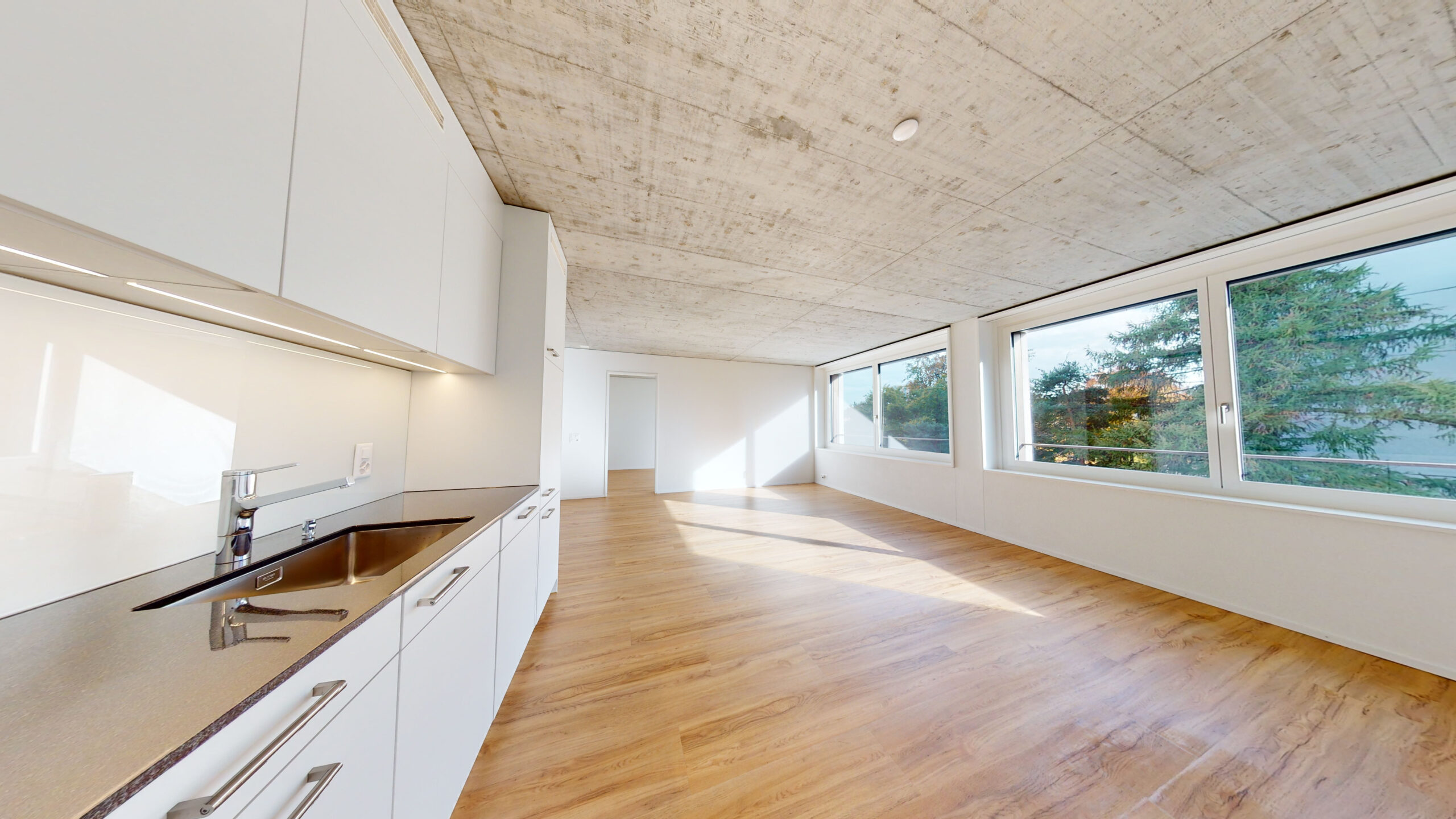 Helles, modernes Apartment-Interieur mit grosszügigem, offenem Grundriss, Holzböden, grossen Fenstern für Tageslicht, einer weissen Küche mit eleganten Schränken und Arbeitsplatten sowie einer Betondecke. Draussen bietet sich ein üppiger, grüner Ausblick.