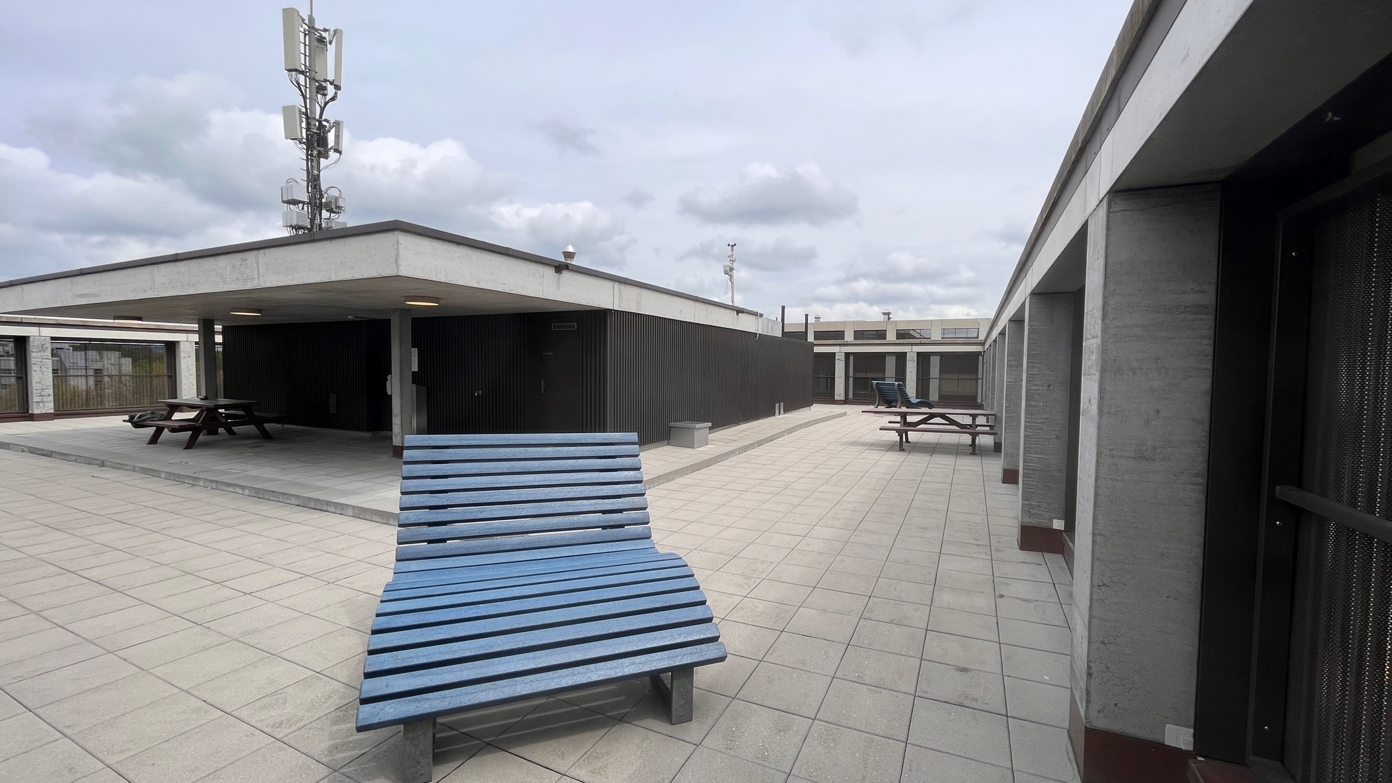 Eine Dachterrasse mit mehreren blauen Holzbänken und Picknicktischen. Der Bereich ist mit hellen Fliesen gepflastert, im Hintergrund sind Mobilfunkmasten auf dem Gebäude zu sehen. Der Himmel ist bewölkt.