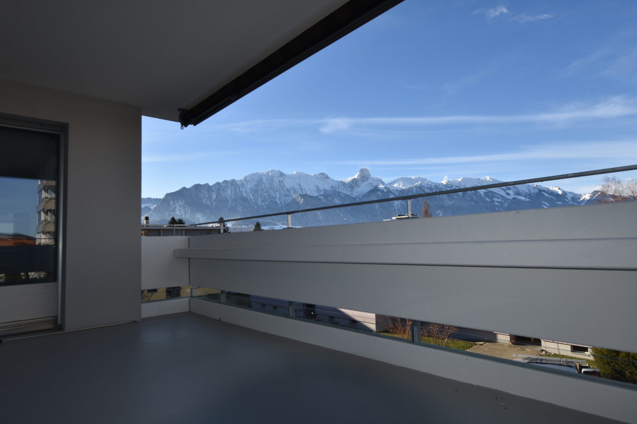 Ein Blick von einem Balkon auf eine schneebedeckte Bergkette unter einem klaren blauen Himmel. Der Balkon hat ein modernes Geländer und am Fusse der Berge sind einige Gebäude zu sehen.