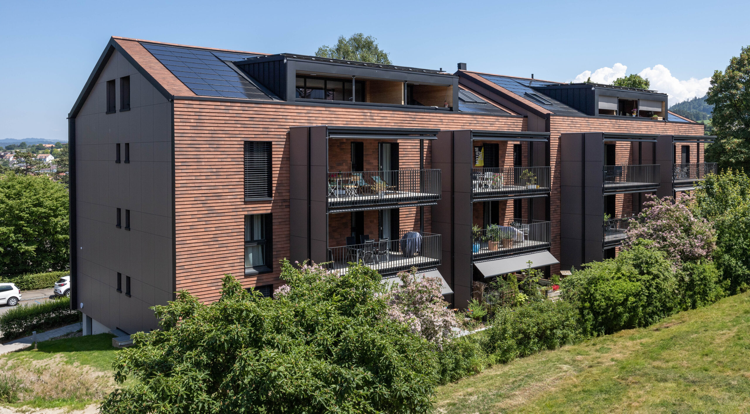 Modernes Mehrfamilienhaus mit Holz- und dunklen Verkleidungen, grossen Fenstern und Balkonen. Auf dem Dach sind Sonnenkollektoren installiert. Umgeben von Grün und Bäumen unter einem klaren blauen Himmel.