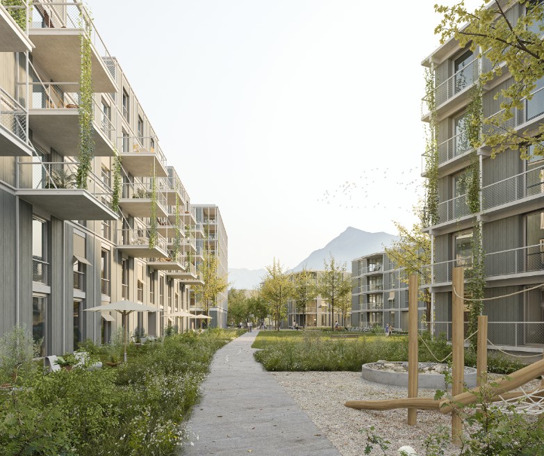 Ein modernes Wohngebiet mit zwei Mehrfamilienhäusern auf beiden Seiten eines von Grün gesäumten Weges. Die Gebäude haben Balkone und Pflanzen ranken an den Fassaden hoch. Im Hintergrund sind Berge unter einem klaren Himmel zu sehen.